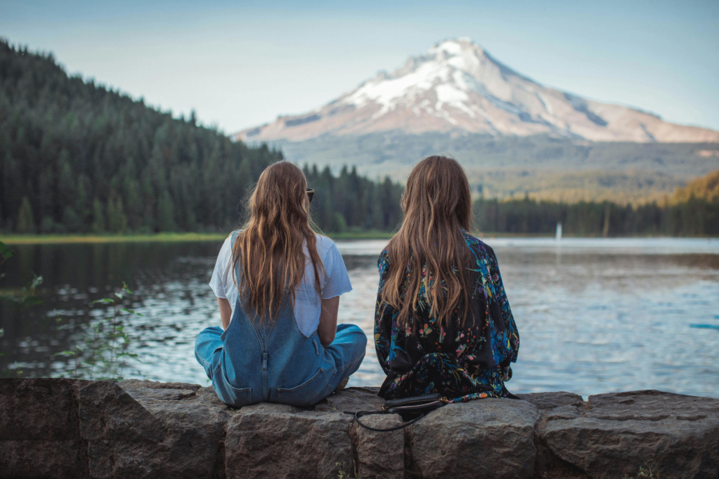 two girls siting on a rock wall in Oregon with river in front of them and the mountains in the background